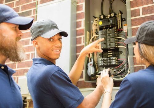 Two men and one woman electricians working on a residential breaker box.  They use tools to assess the repair and wear matching blue uniforms.  The multi-ethnic group is discussing next steps in the job repair.  Electric meter to side.